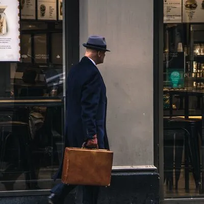Picture of a man in a suit jacket and blue hat with brown leather gloves and a leather brief case walking away down a London street.
