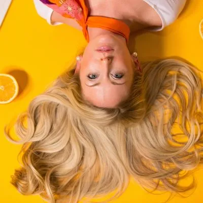 Blonde women laying on a orange surface with half cut oranges around her.