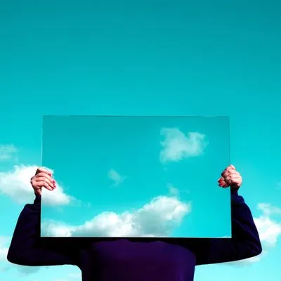 Person holding up a mirror to cover their face and reflect back a blue cloudy sky.